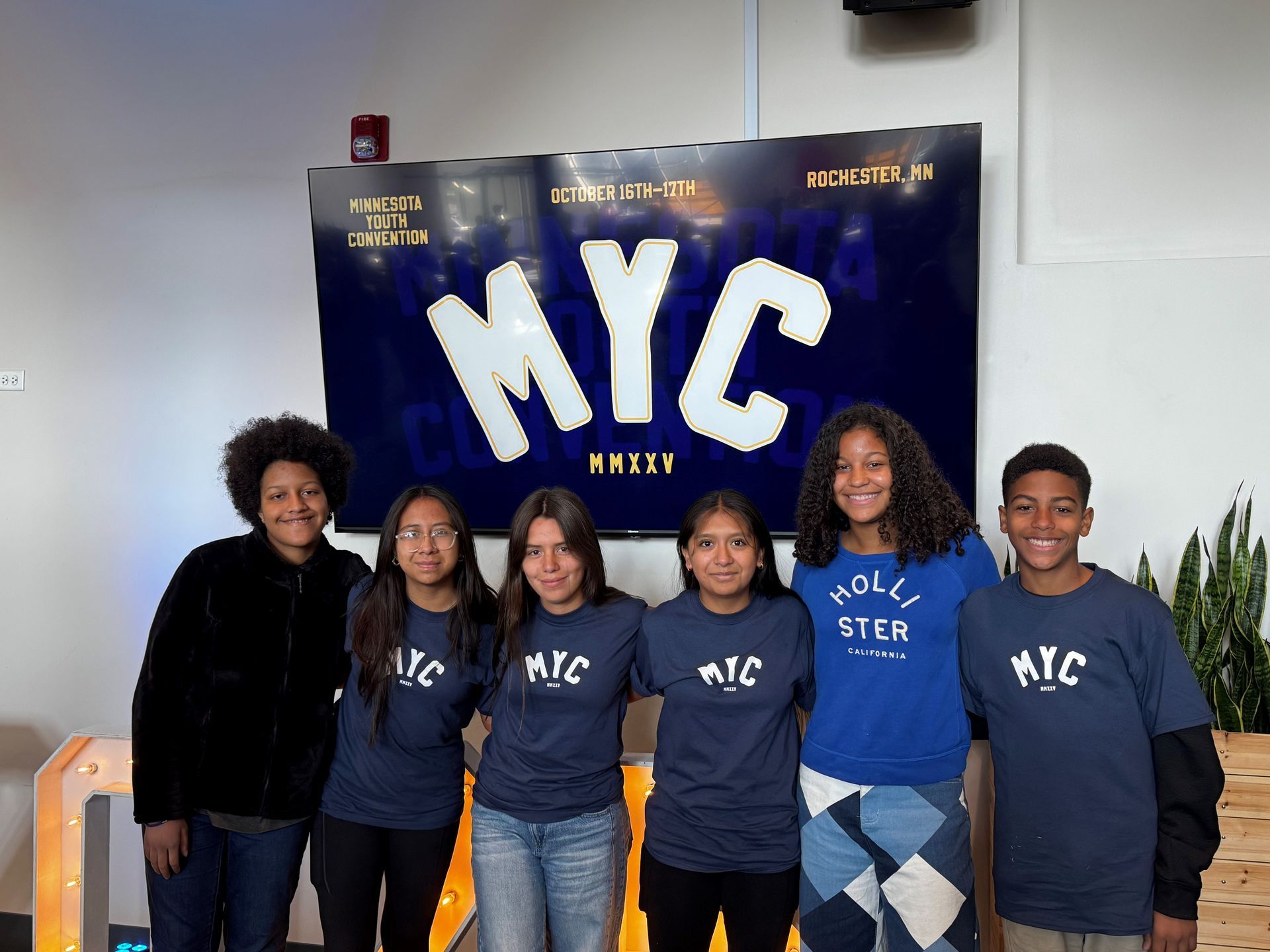 Five people posing indoors in front of a blue “MYC” backdrop, wearing matching shirts.
