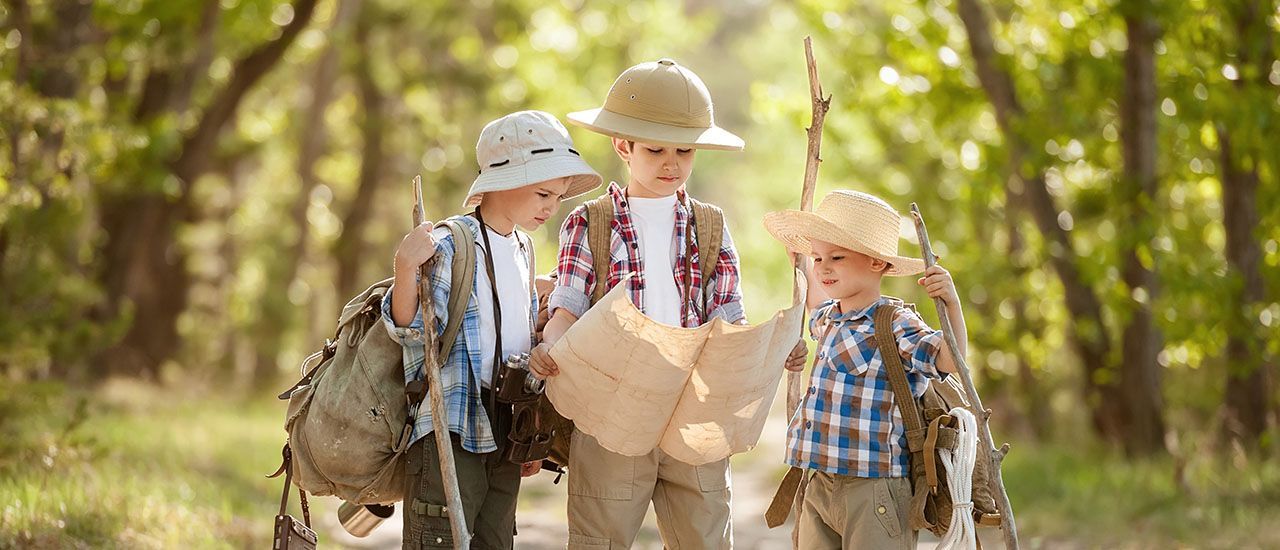 Three children dressed as explorers wearing hats and backpacks stand in a sunny forest studying a paper map.