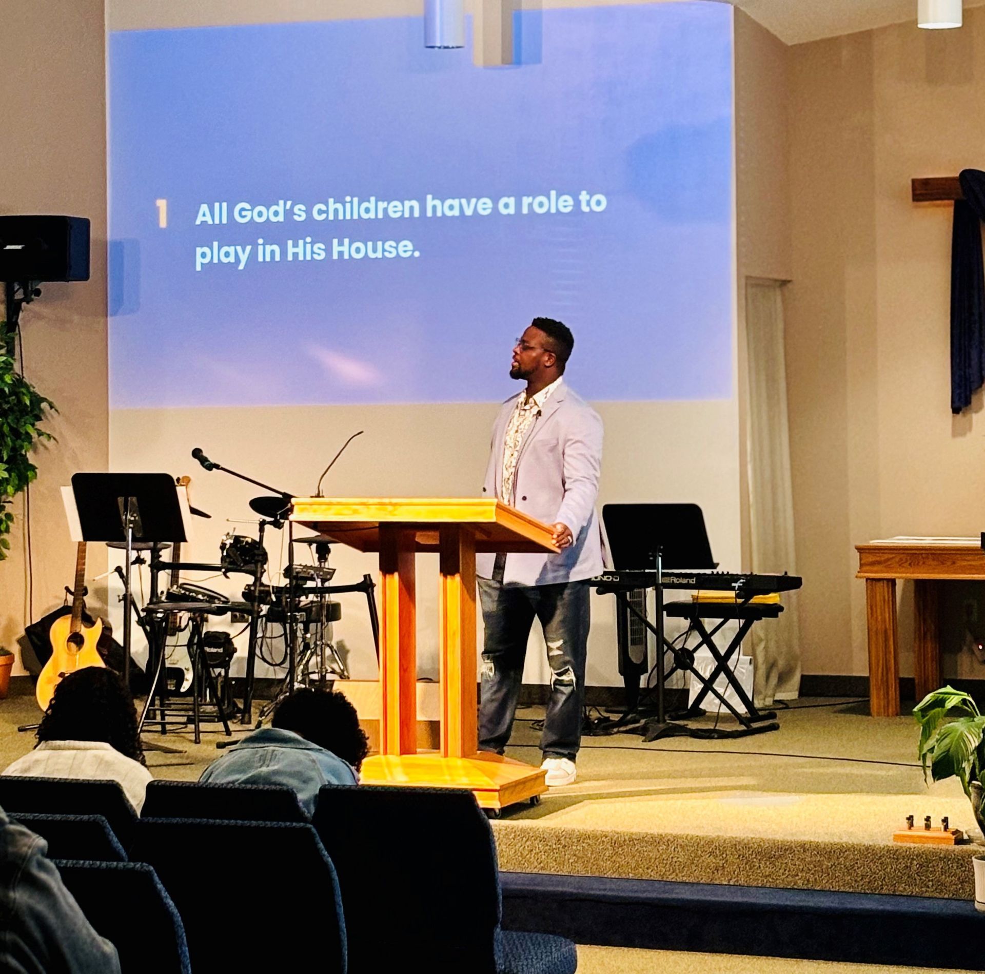 Speaker at church lectern on stage with projected slide reading, “All God’s children have a role to play in His House.”