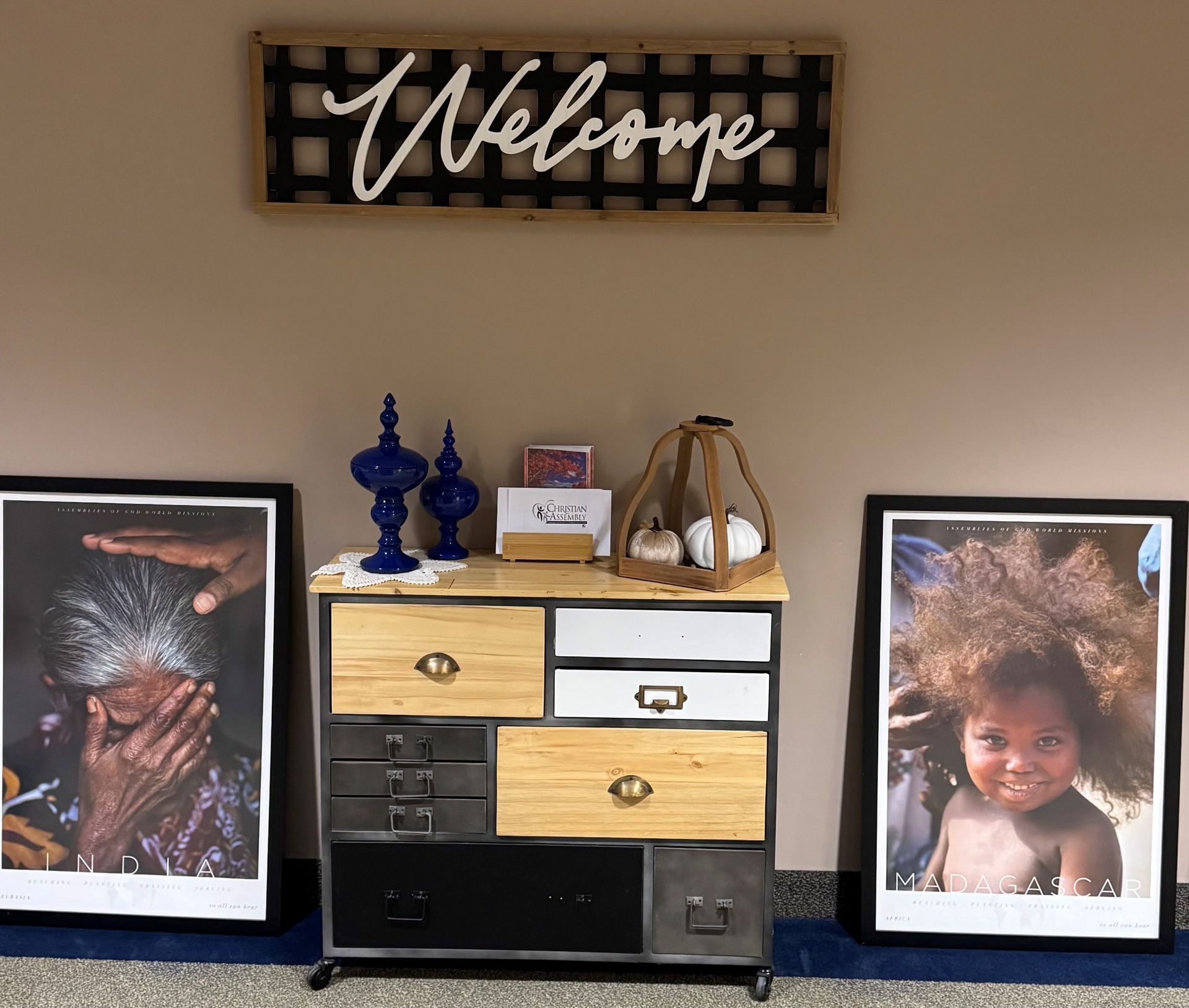 Wall display with a “Welcome” sign above a dresser holding decor and two framed photos on either side