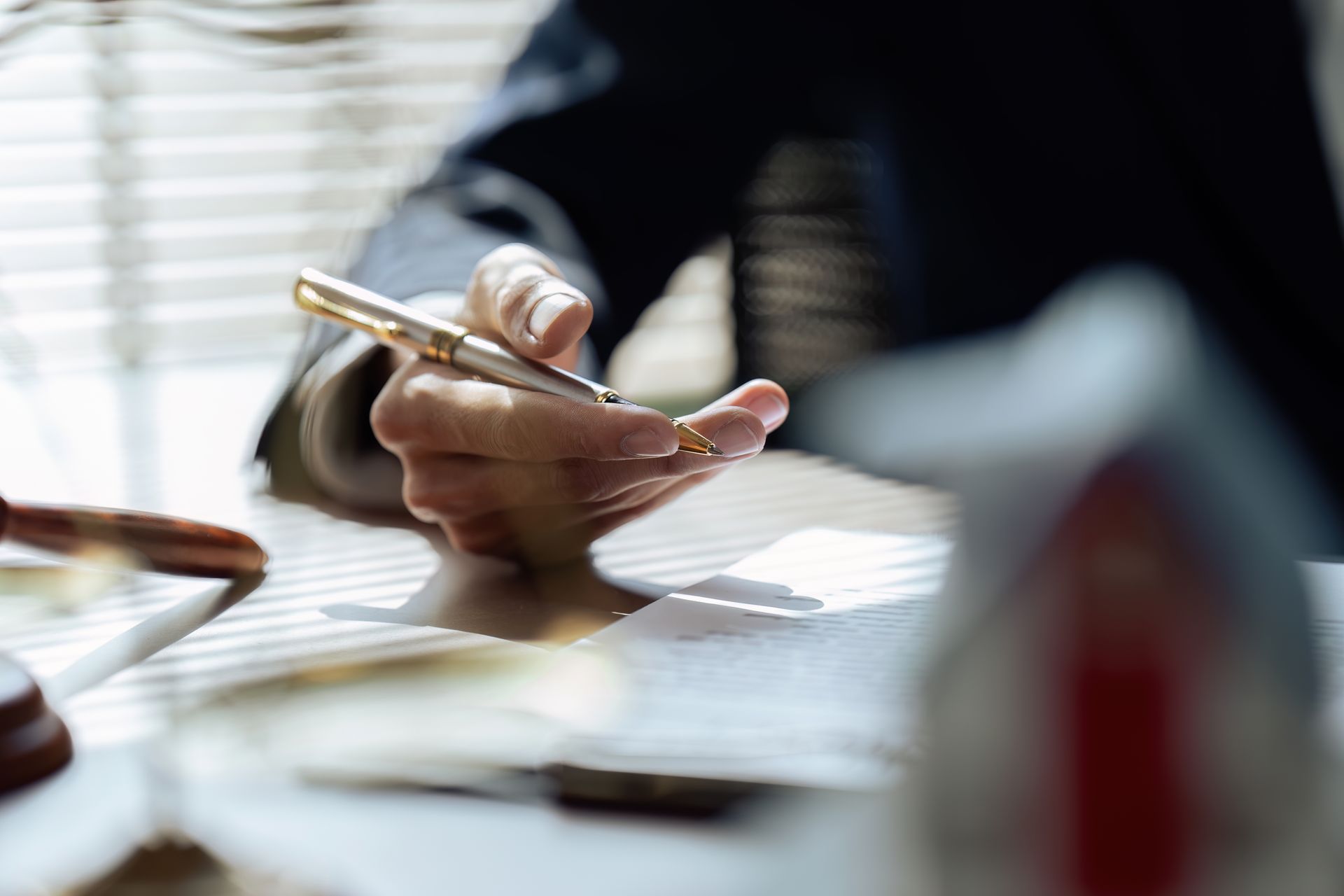 Person's hand holding a pen over papers on a desk; window in the background.