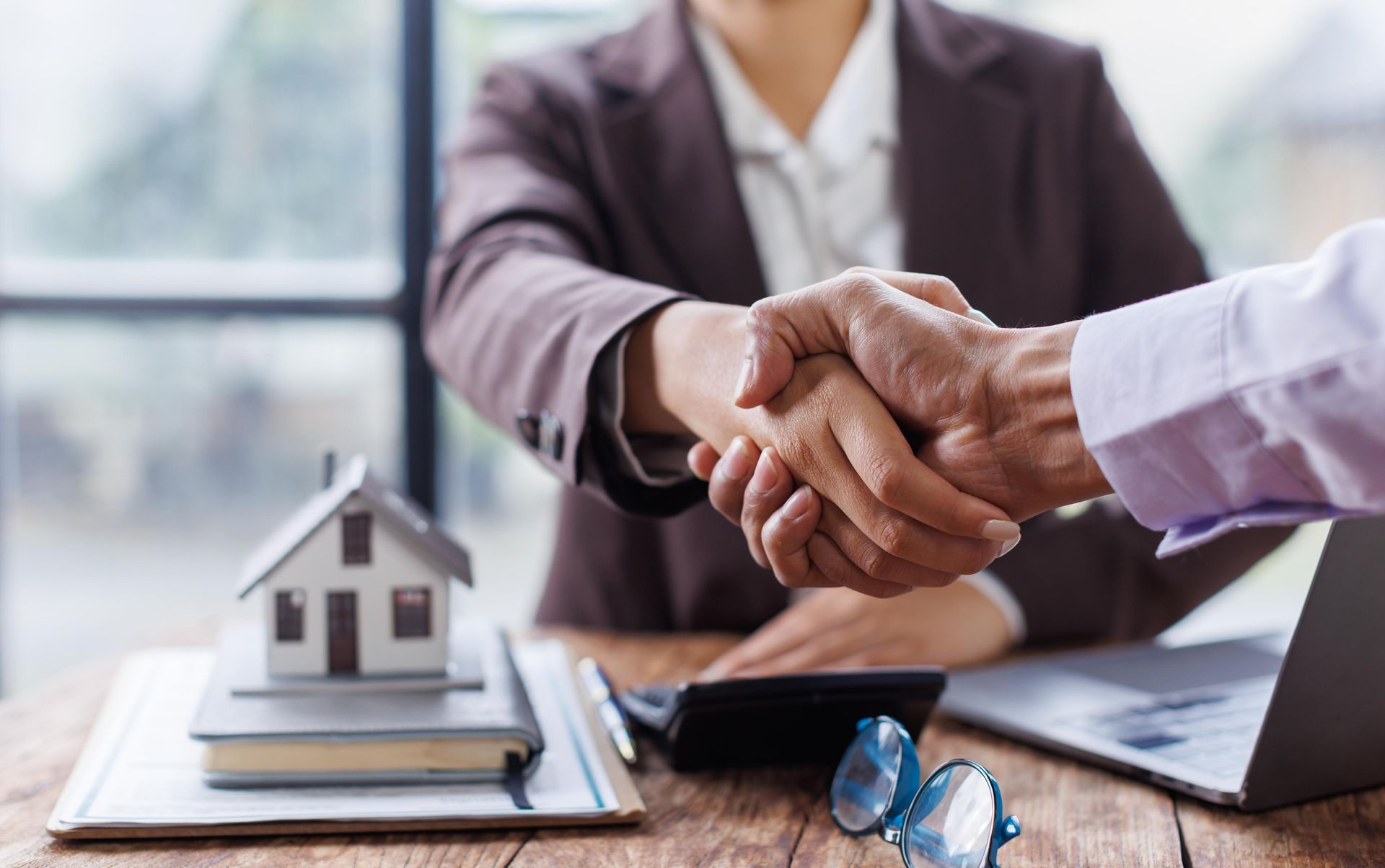 People shaking hands, miniature house, laptop, and documents on a desk, possibly a real estate transaction.