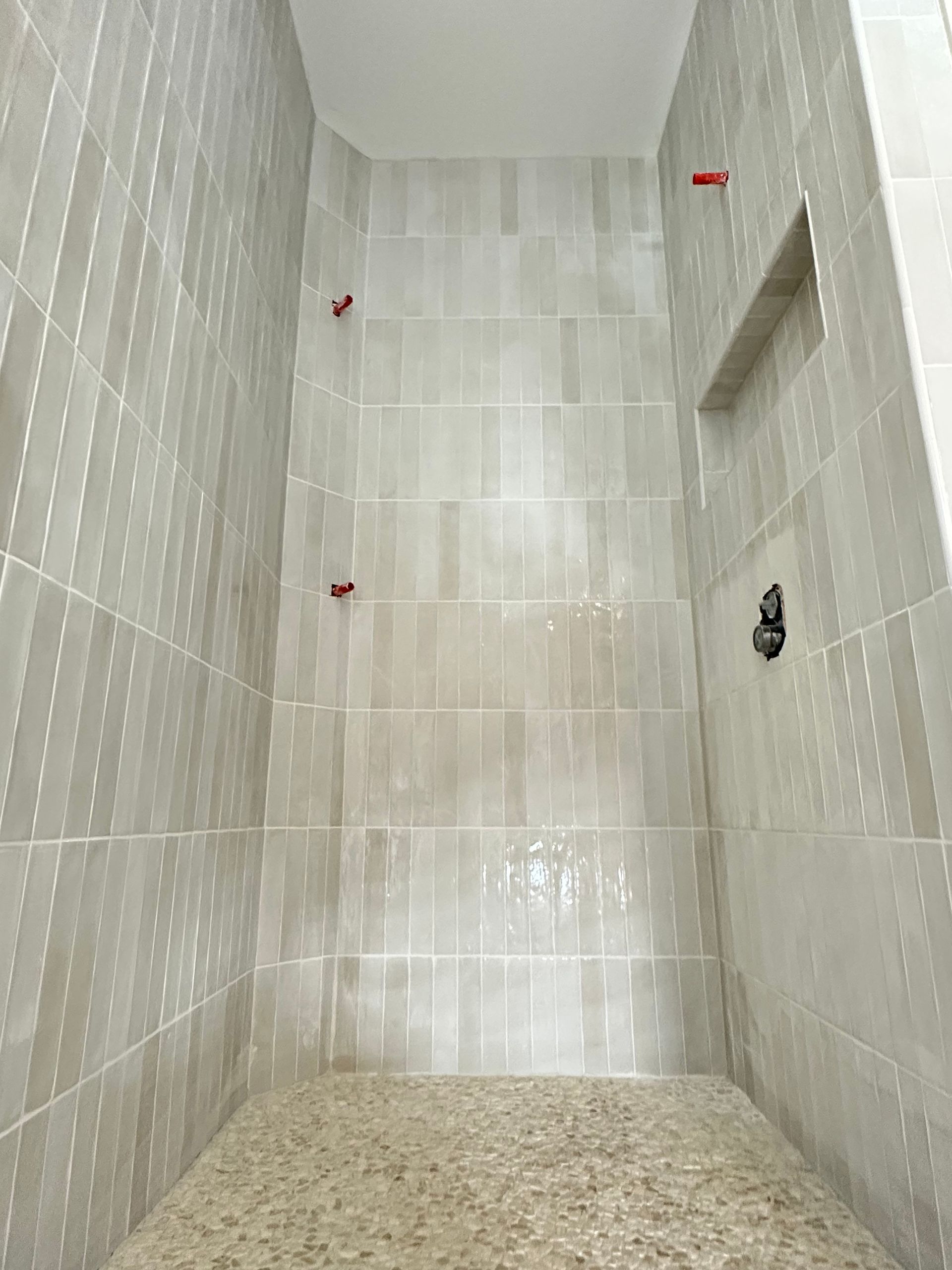 Narrow tiled shower stall with beige walls, pebble floor, and red wall fixtures
