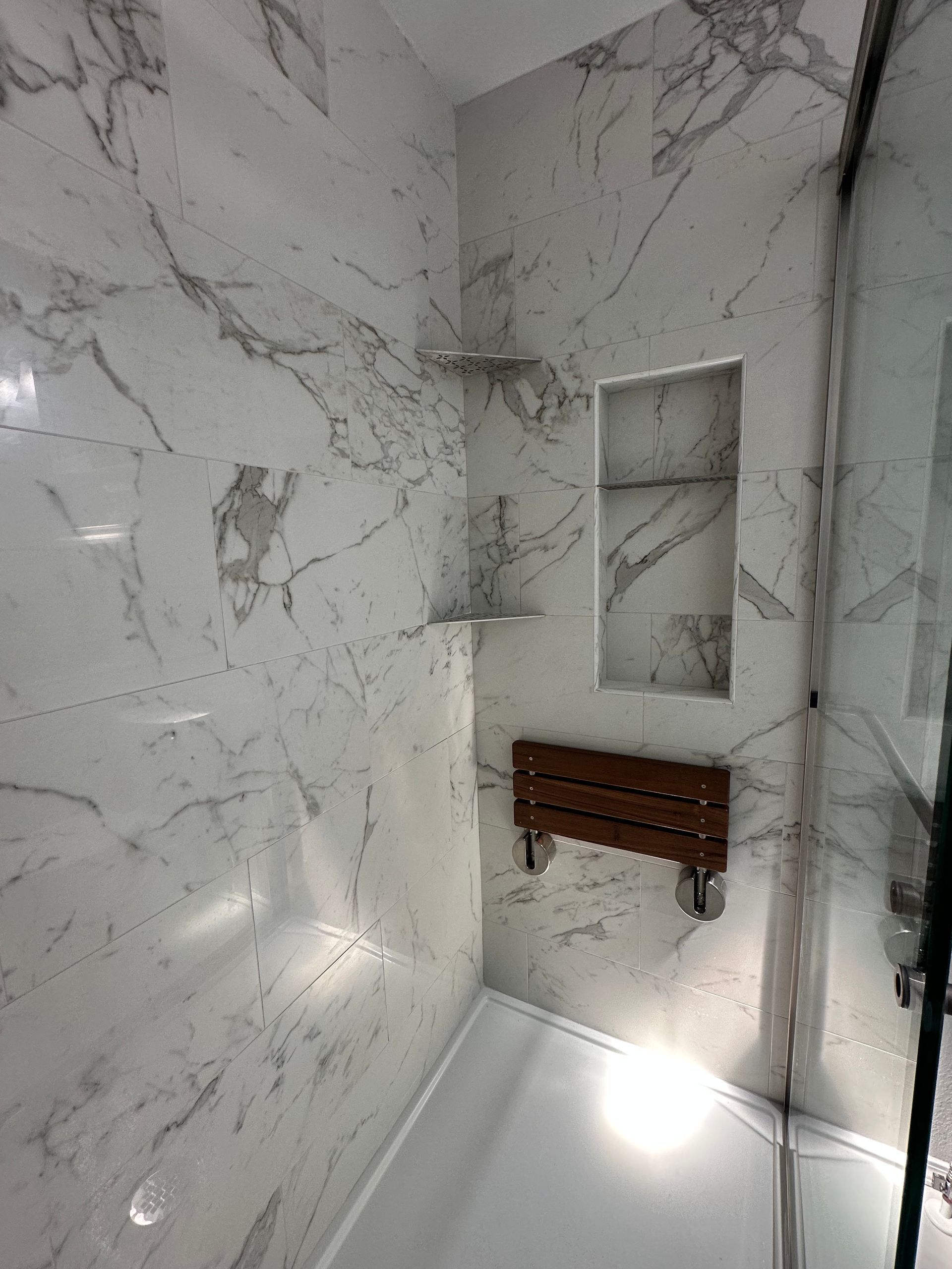 Marble-tiled shower corner with built-in shelves and a wooden bench under bright light
