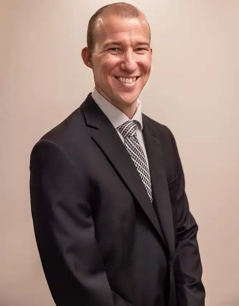 Smiling man in a dark suit and striped tie against a plain beige background
