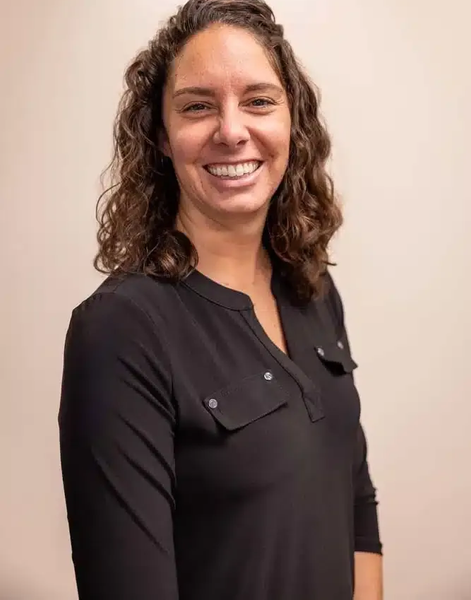 Smiling person in a black blouse standing against a light beige background.