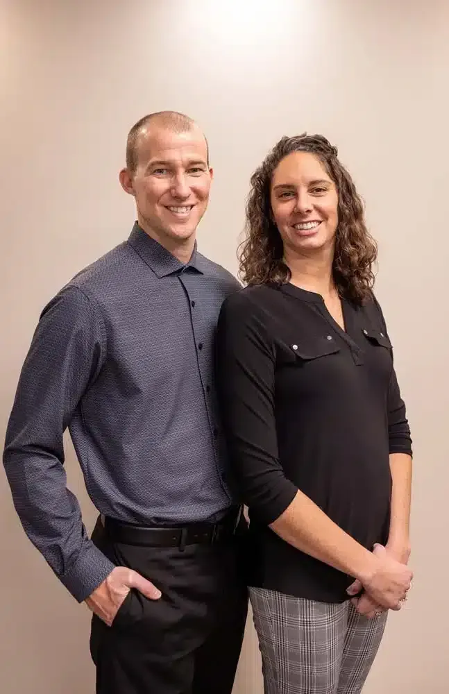 Two people standing indoors, smiling, in business attire against a plain wall.