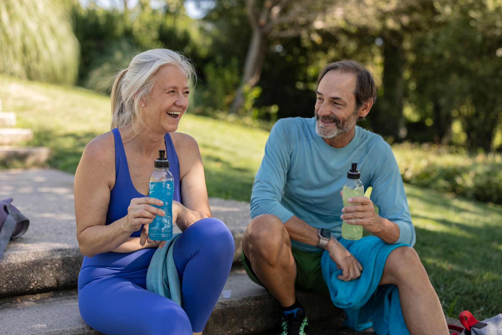 Two people sitting outdoors, smiling and holding water bottles on a sunny hillside.