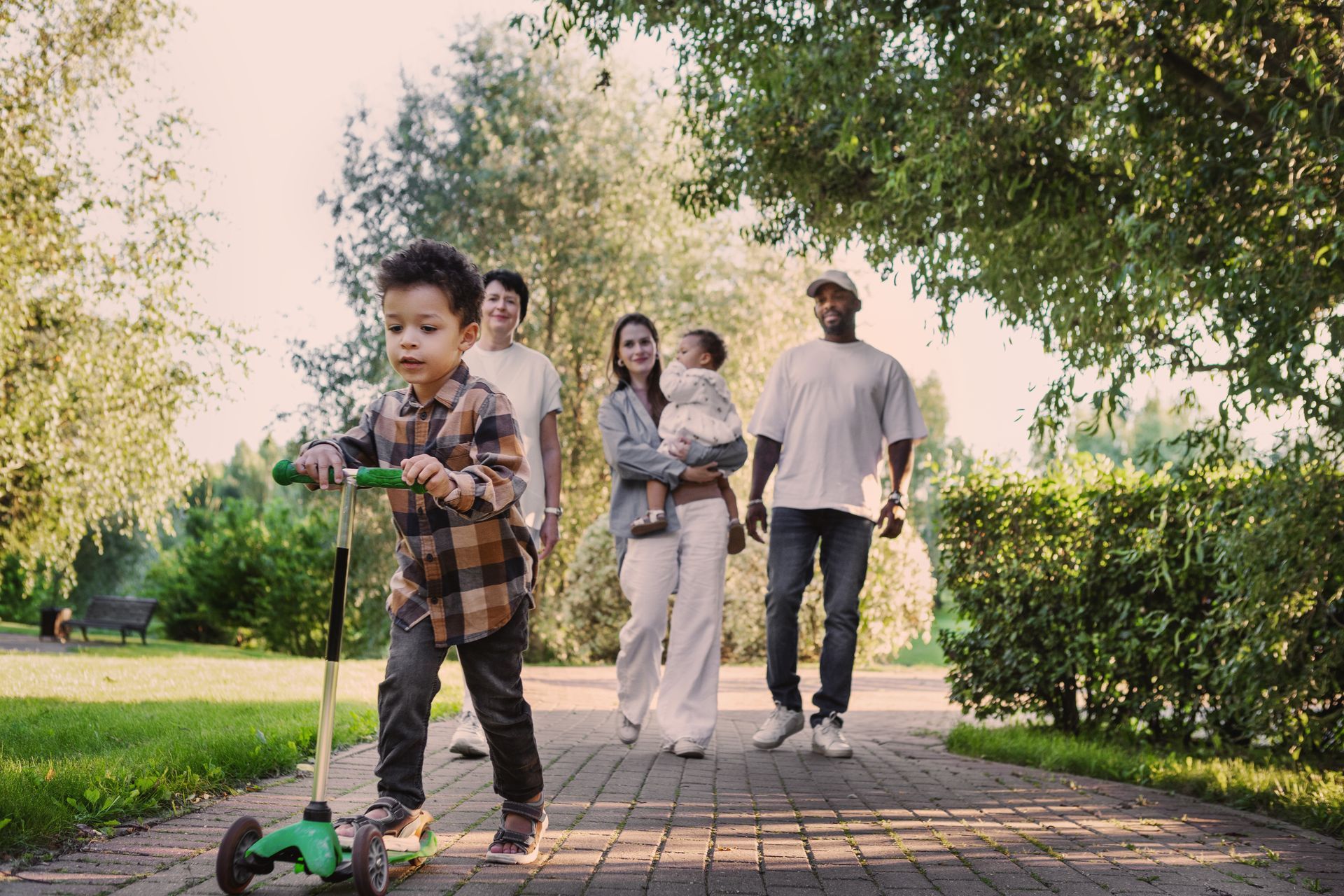 Child riding a scooter on a brick path while adults walk behind in a sunny park