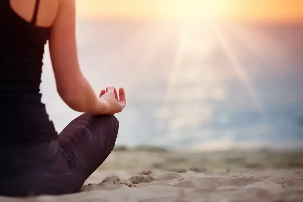 Person meditating on a sandy beach at sunset, seated cross-legged with warm light over the water