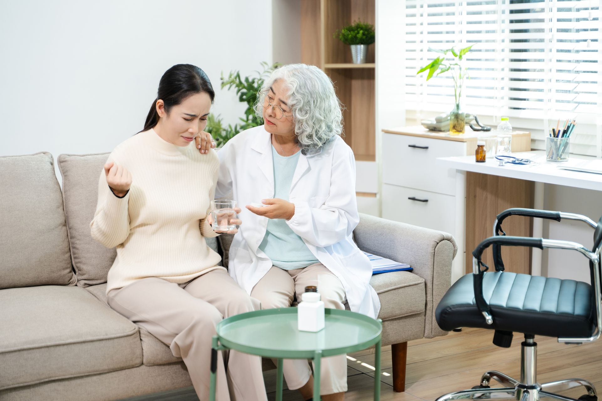 Two women talking on a sofa in a bright living room, with a walker and chair nearby.