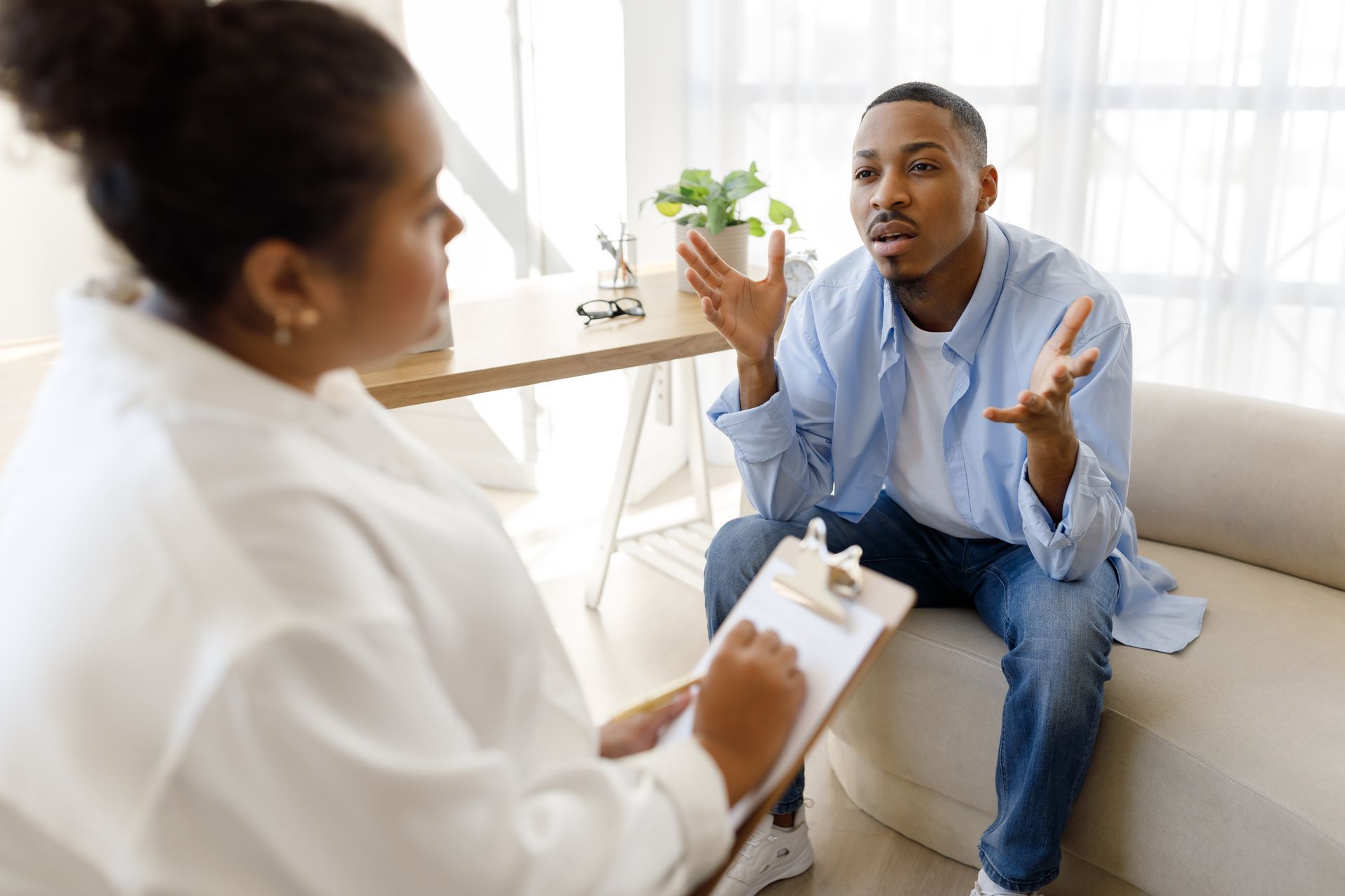 Therapist talking with a patient in a bright office, holding a clipboard while the patient gestures seated on a couch
