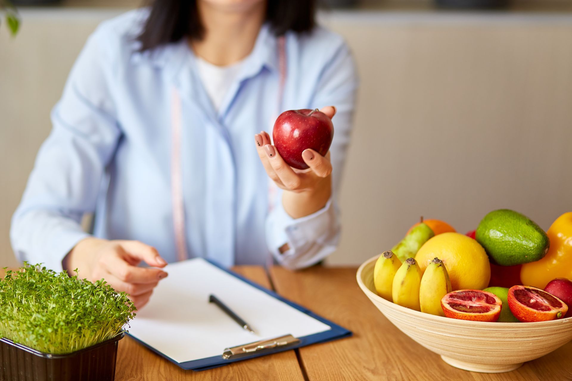 Person holding a red apple at a desk with a clipboard, fruits in a bowl, and green sprouts nearby