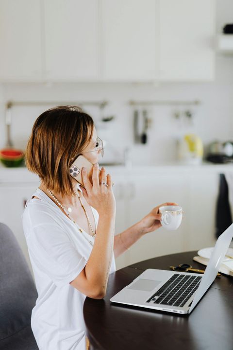 Woman on phone beside open laptop, holding a mug in a bright kitchen