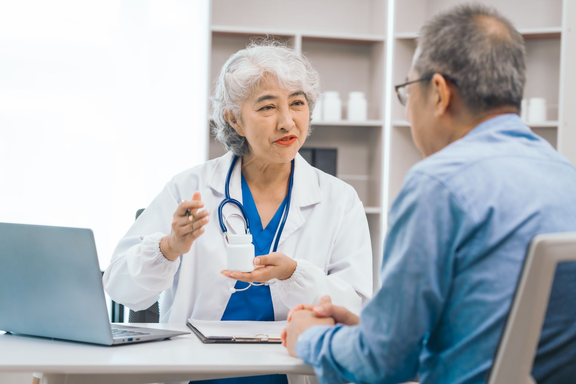 Doctor speaking with a patient in a clinic office, holding a pen beside an open laptop.