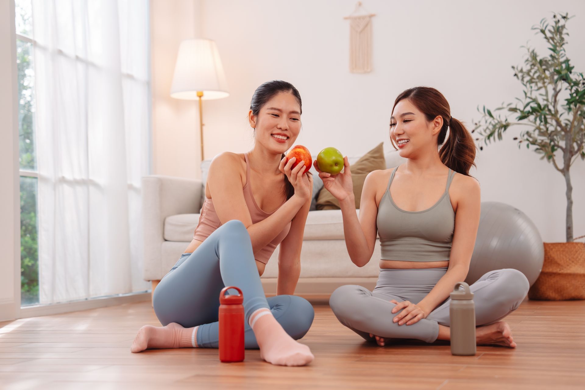 Two women sitting on yoga mats, smiling and clinking fruit in a bright living room