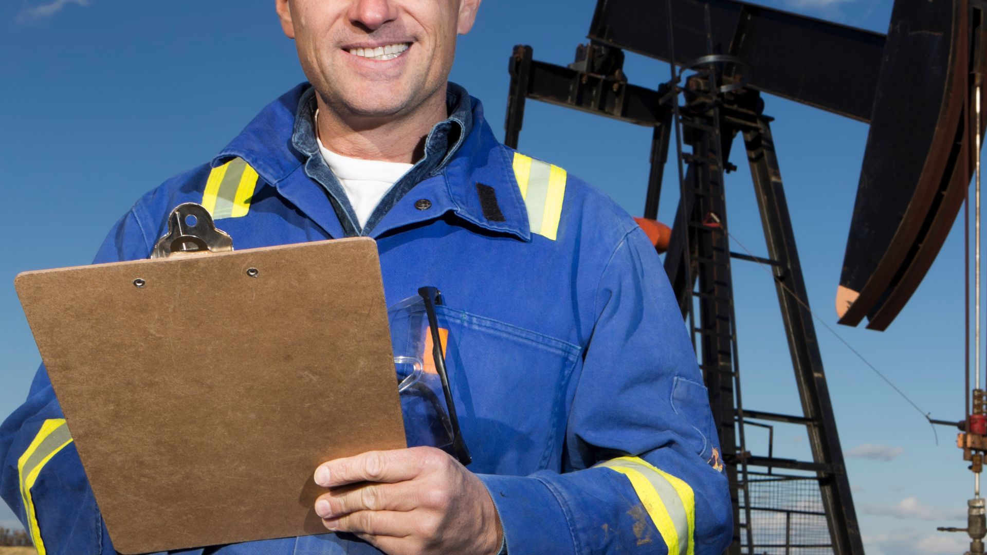 A man in a blue jacket is holding a clipboard in front of an oil pump.