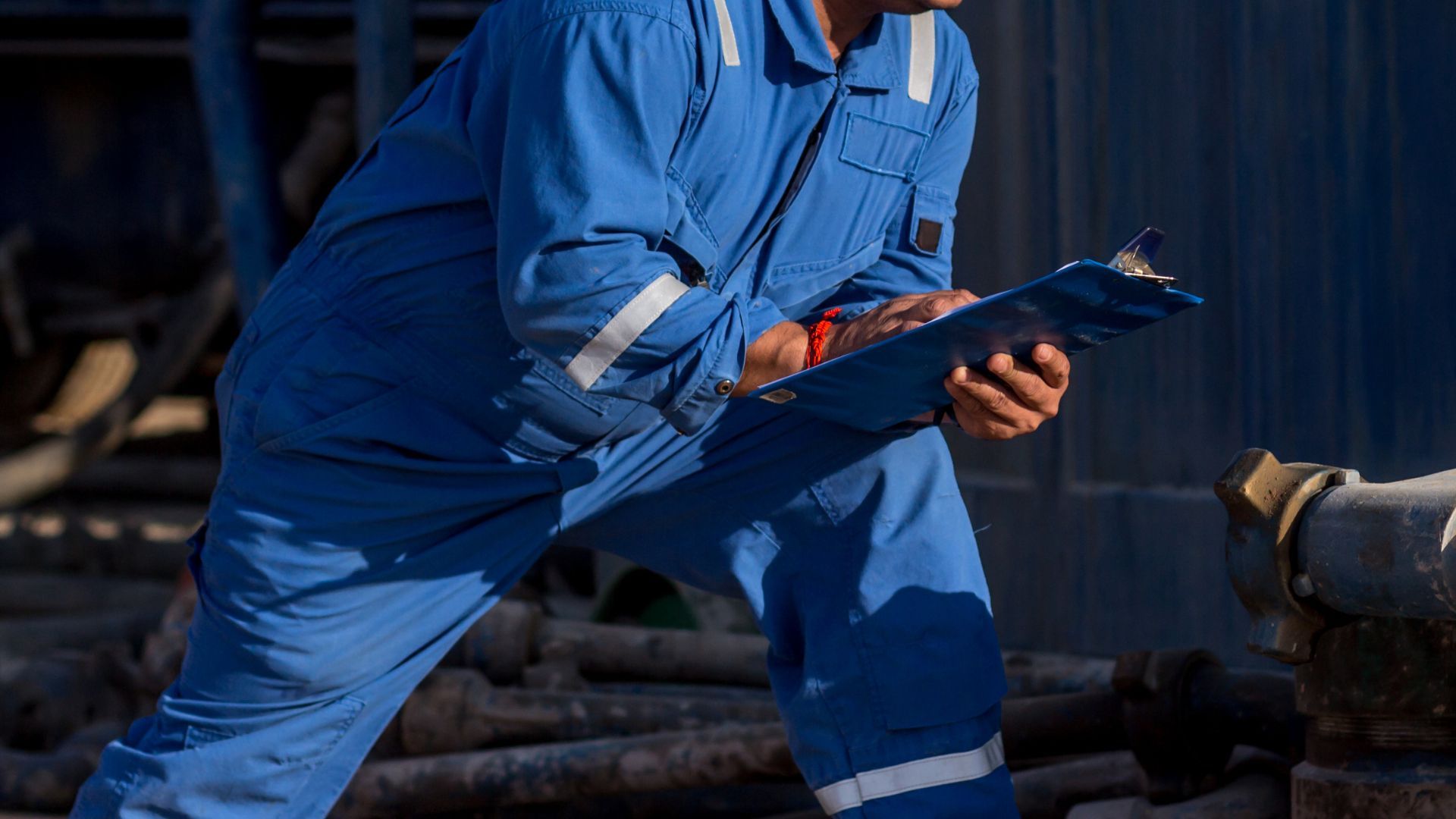 A man in a blue jumpsuit is holding a clipboard and writing on it.