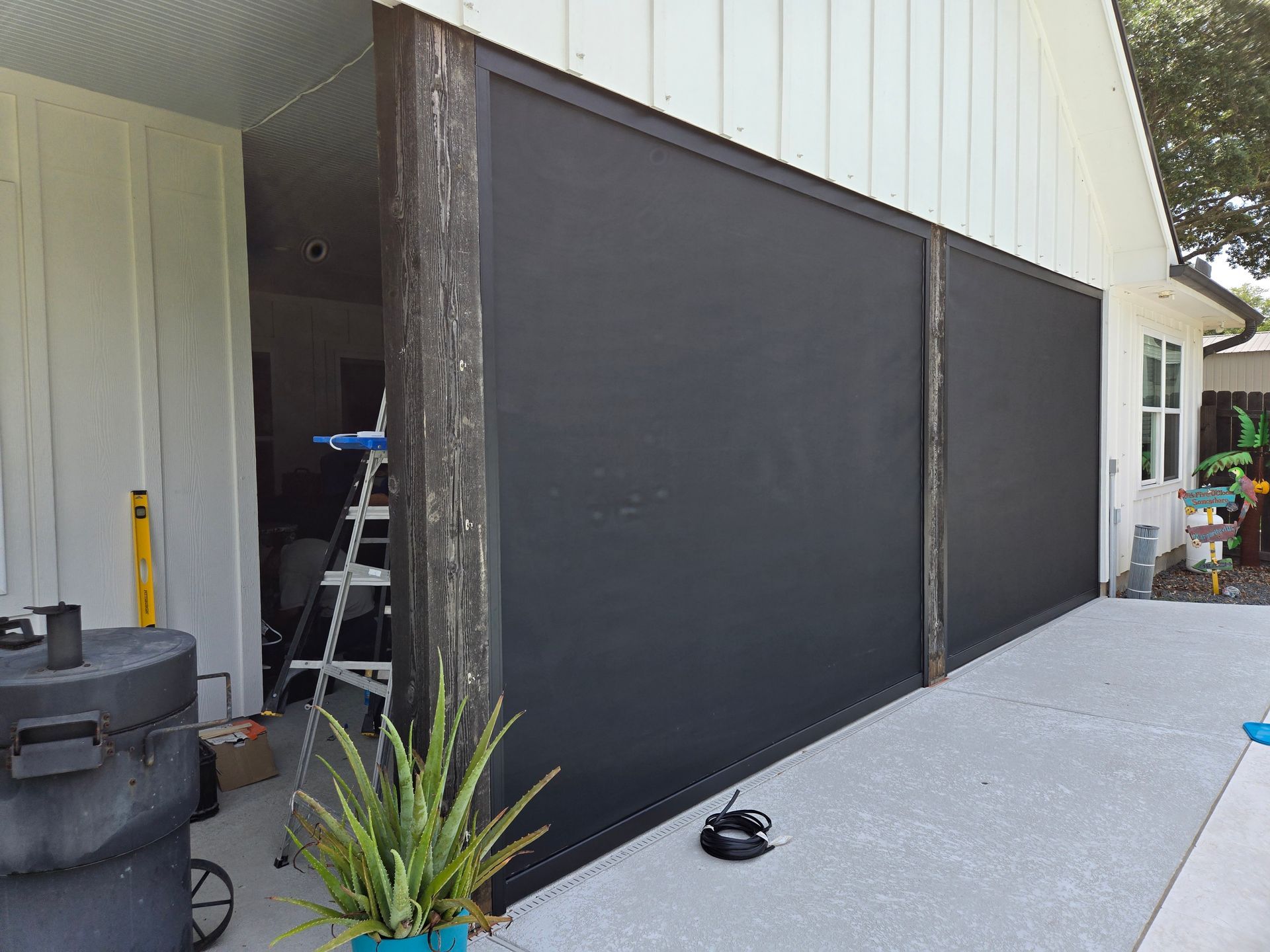 Black privacy screens beside a house on a concrete patio, with a potted plant and tools nearby