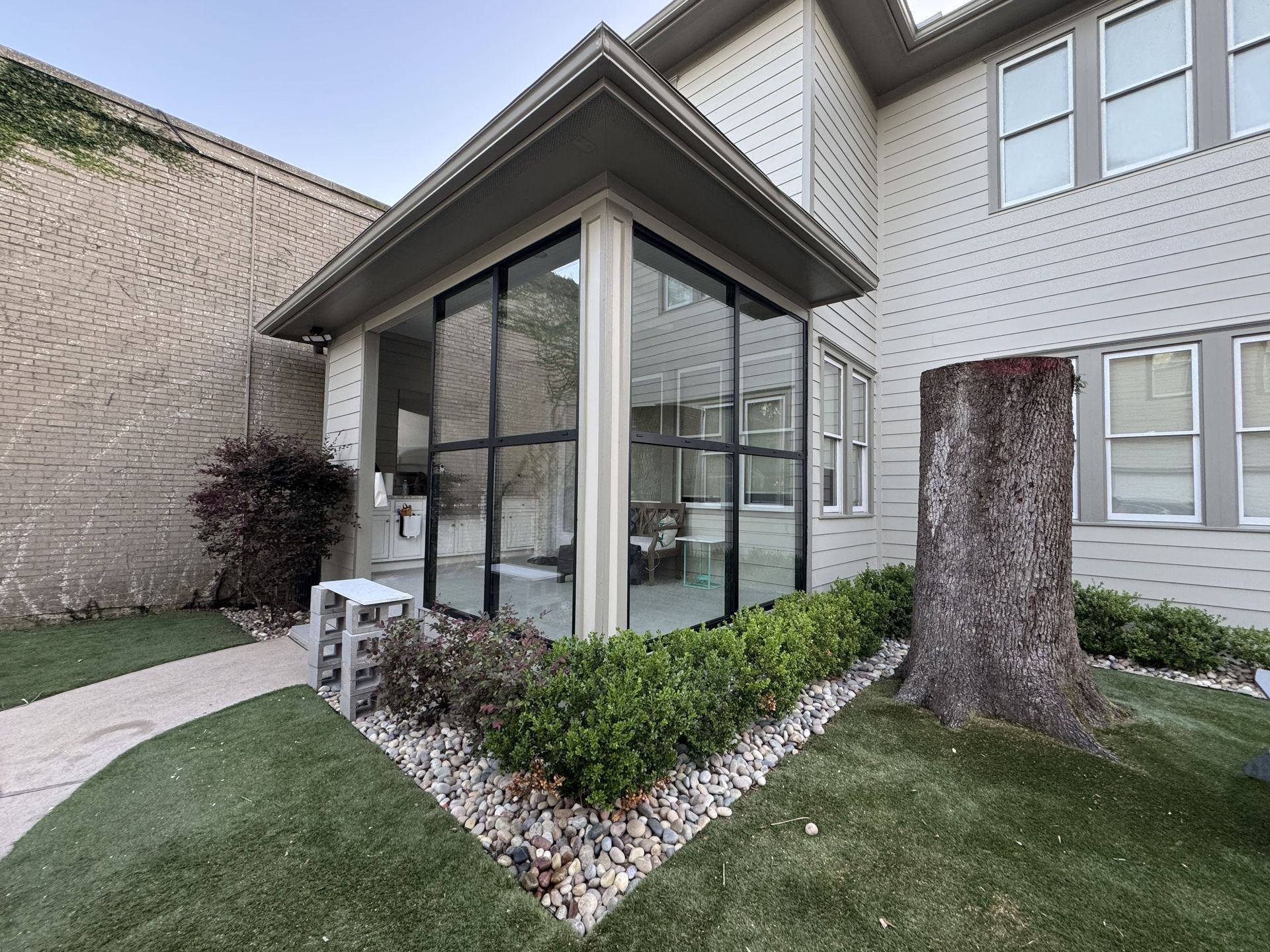 Glass-enclosed patio entrance beside a townhouse, with white siding, shrubs, and a tree stump in the foreground