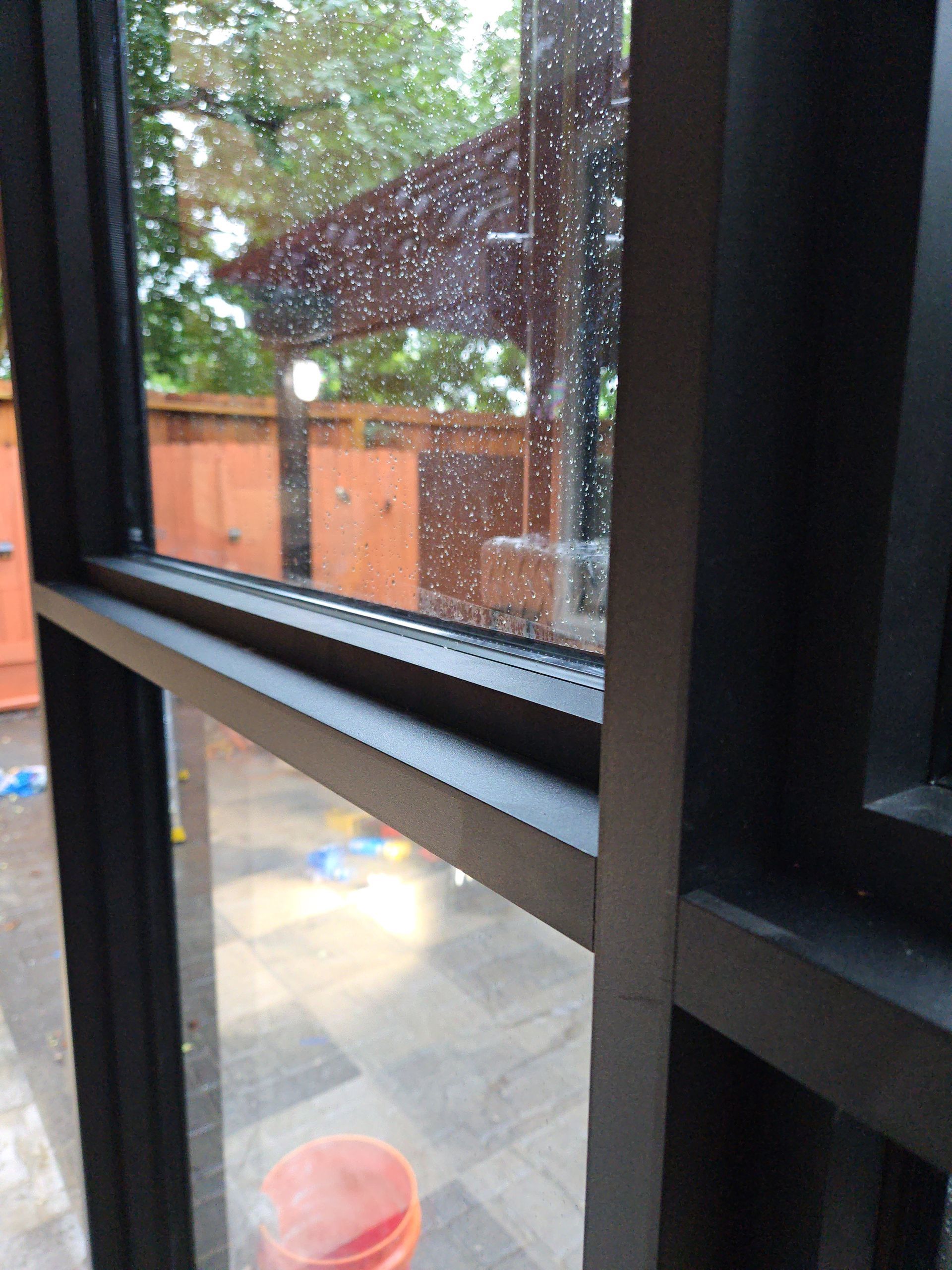 View through a rain-speckled window of a patio with orange shed, trees, and a red bucket below