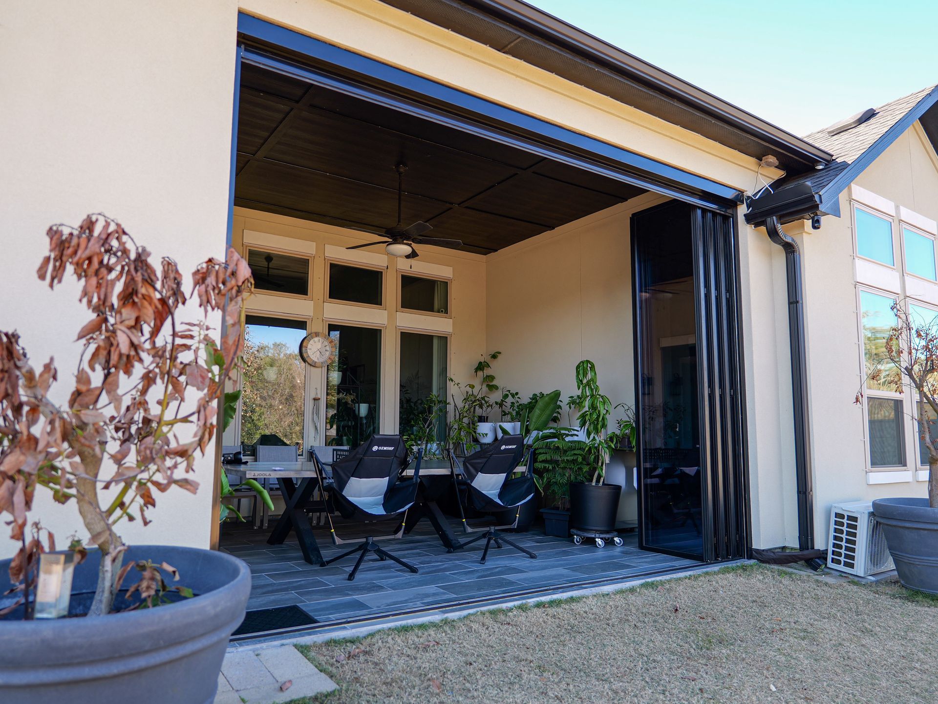 Patio with black chairs and plants beside a light-colored house, viewed from the garden