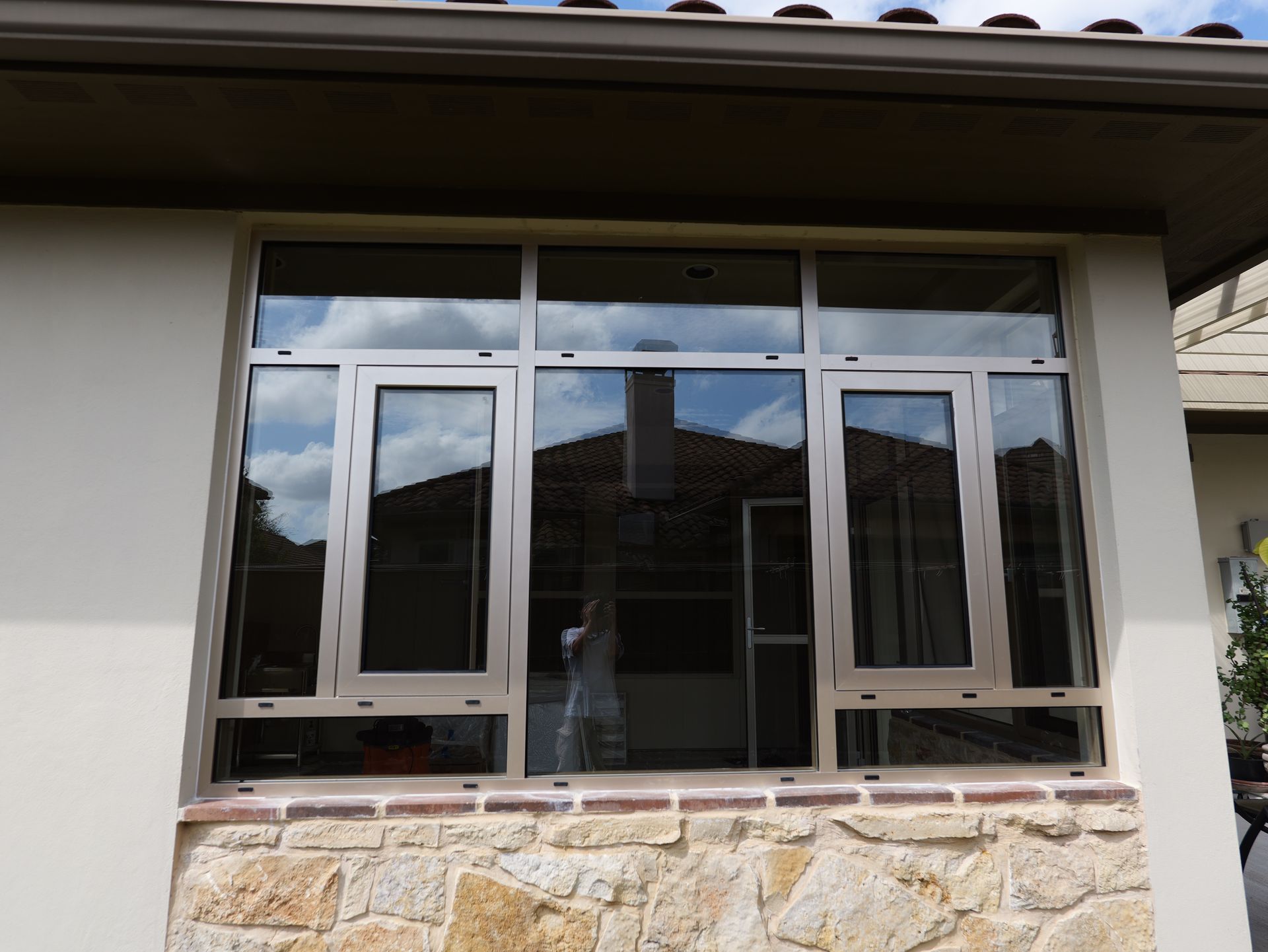 Front view of a house window with white frames, reflecting clouds and a roof, above a stone wall.