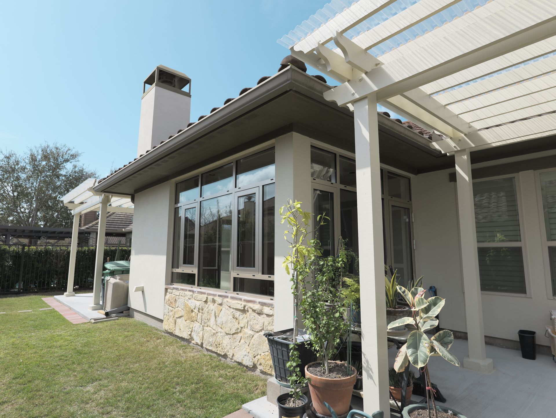 Patio with white pergola, stone planter, and large windows beside a house in a sunny yard