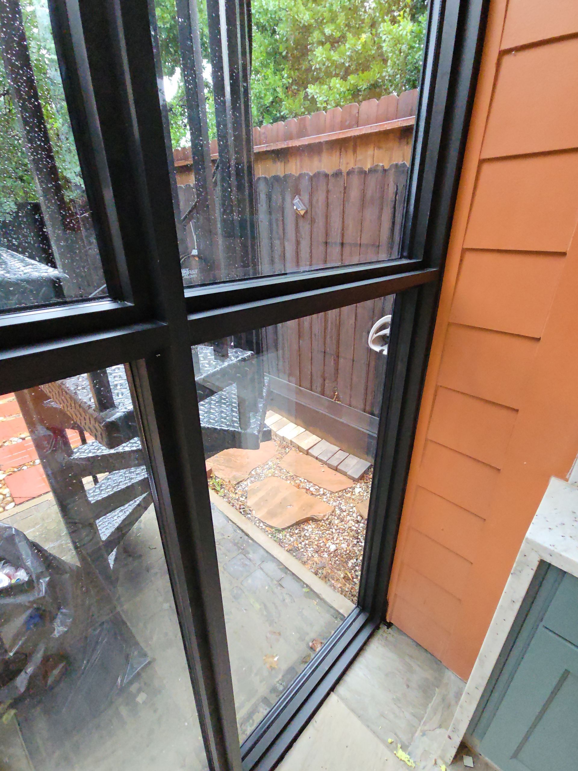 View through a black-framed window to a small fenced patio with stone pavers and a wooden chair.