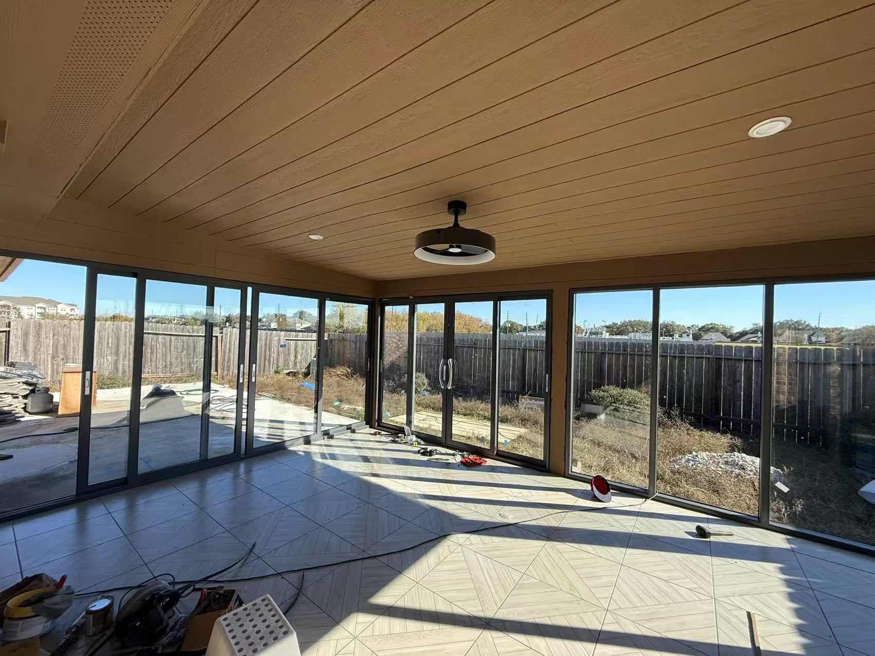 Covered patio with glass walls, concrete floor, and a fan, overlooking a fenced yard in daylight