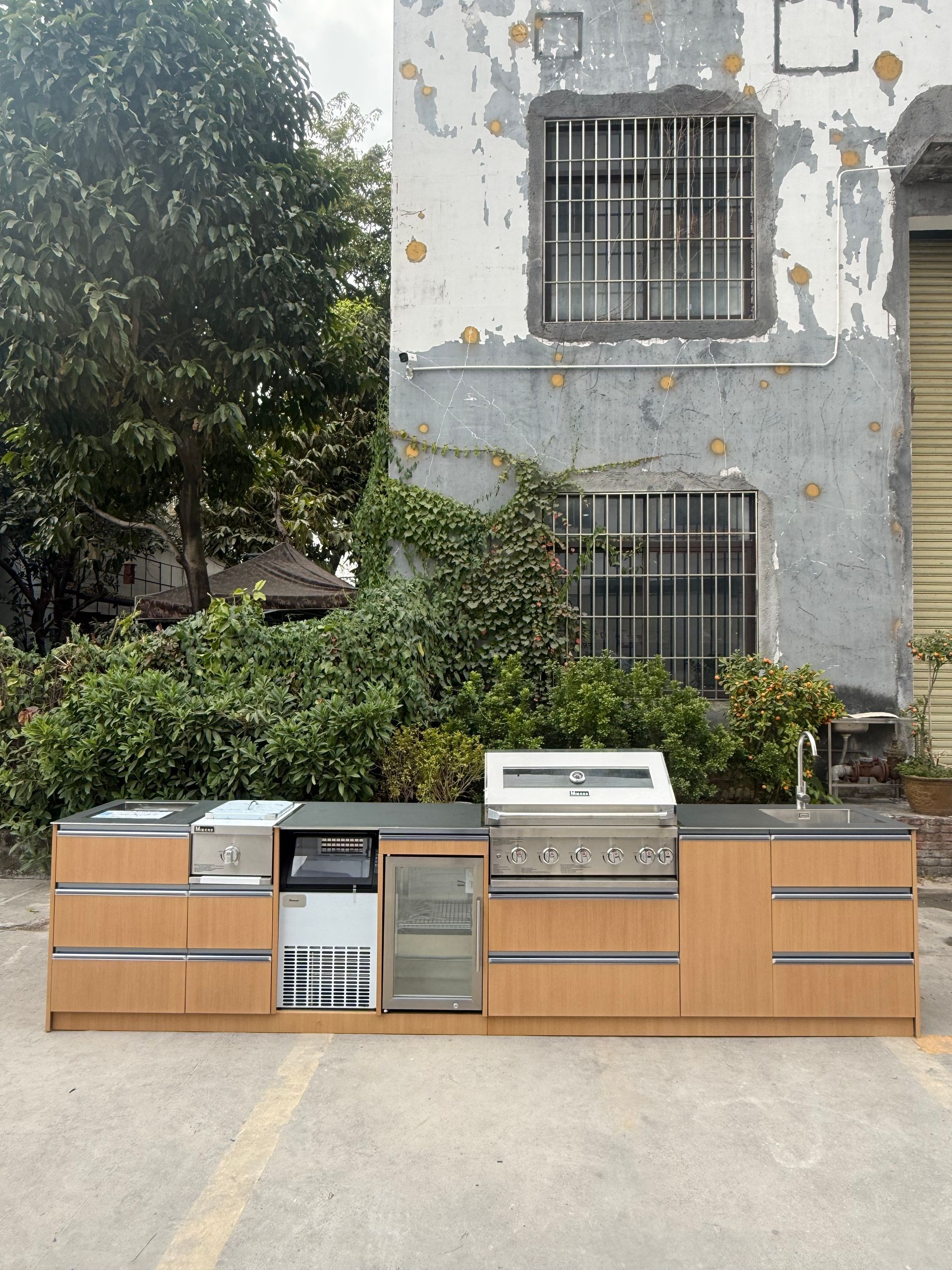 Outdoor kitchen setup with wood cabinets, stainless appliances, and a grill against a building wall.