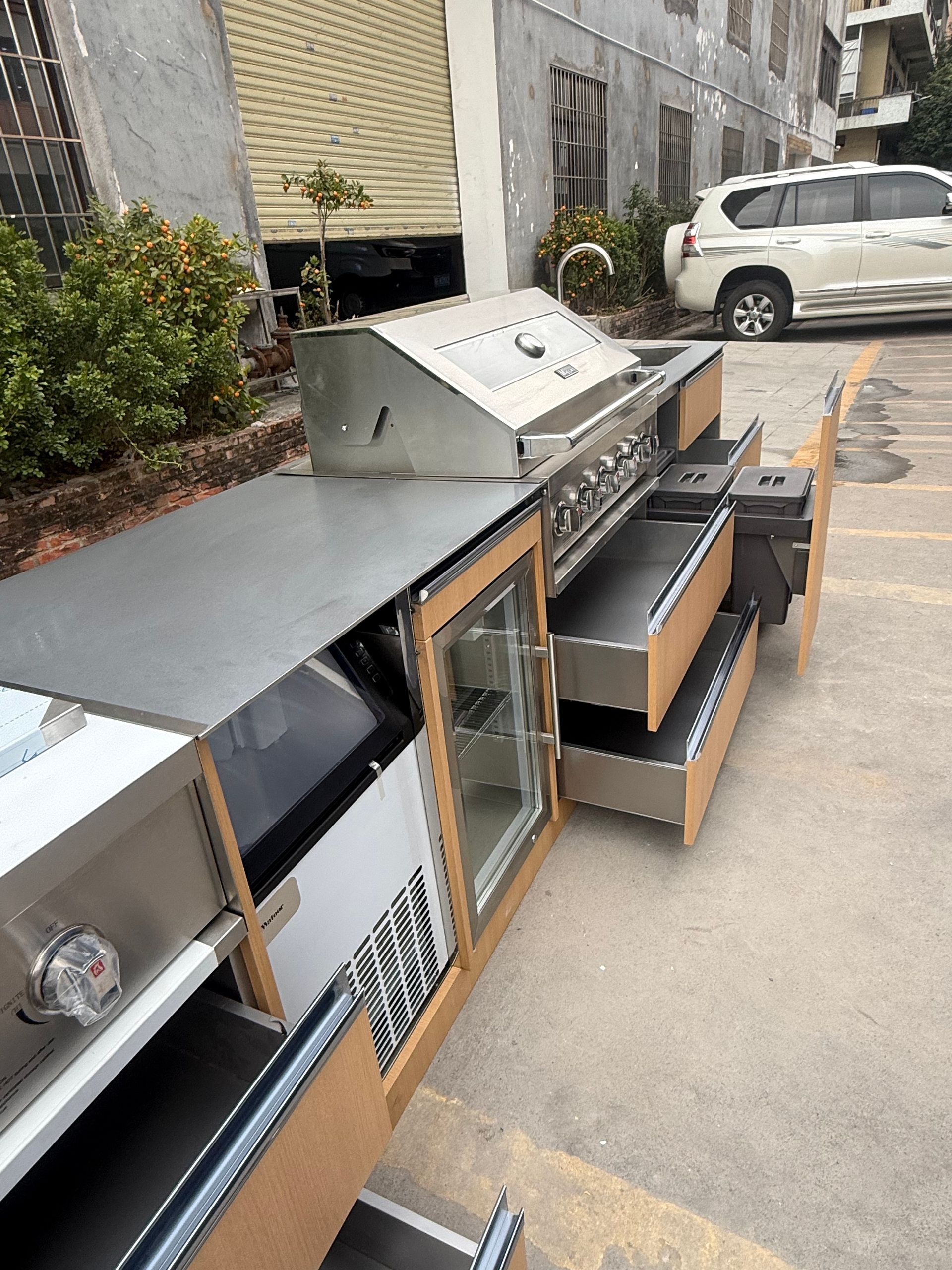 Outdoor row of stainless steel kitchen appliances and cabinets on a sidewalk near parked cars.