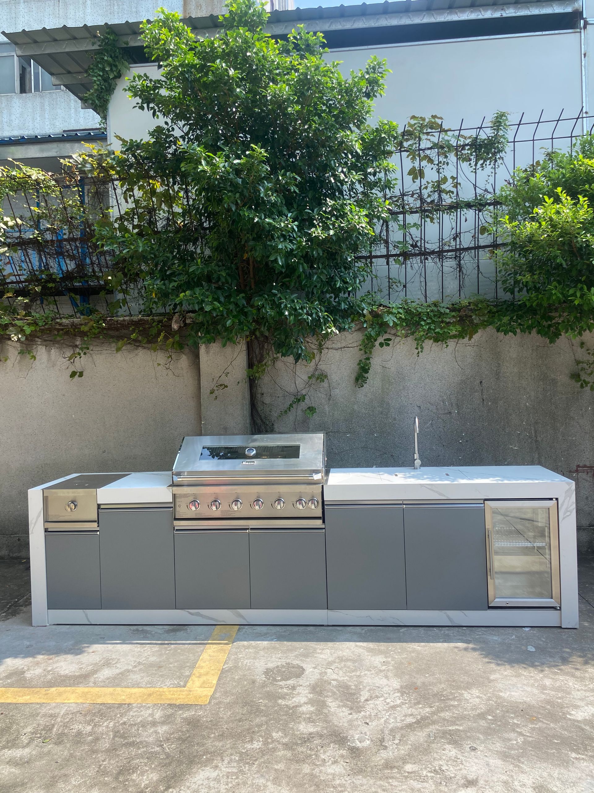 Outdoor stainless steel grill cabinet against a concrete wall with greenery behind it.