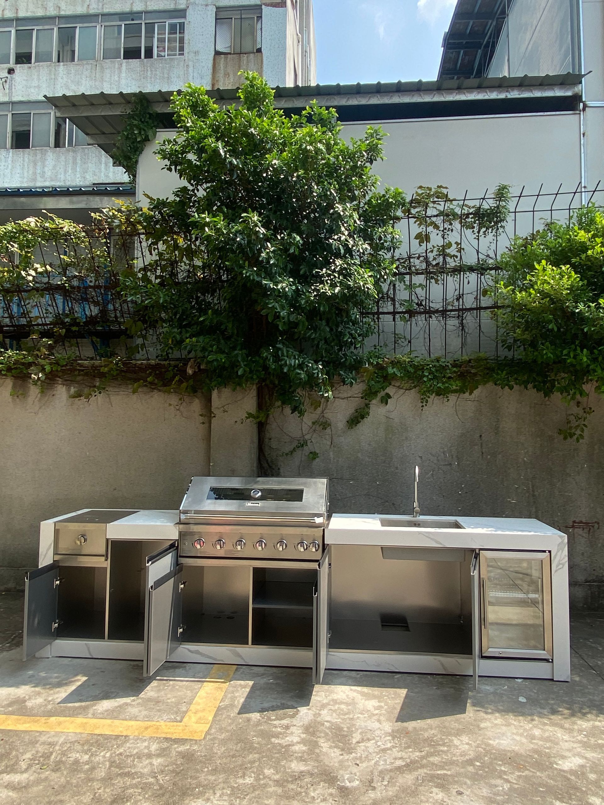Outdoor metal sink and cabinet units against a wall, with a grill in the center and a small tree behind them.