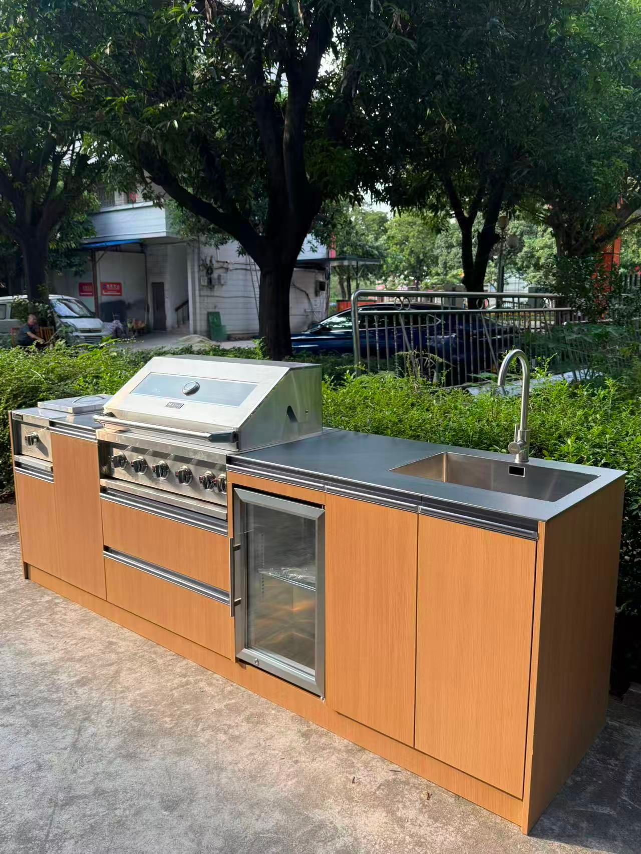 Outdoor stainless-steel grill island with wood cabinets and a sink in a park-like setting
