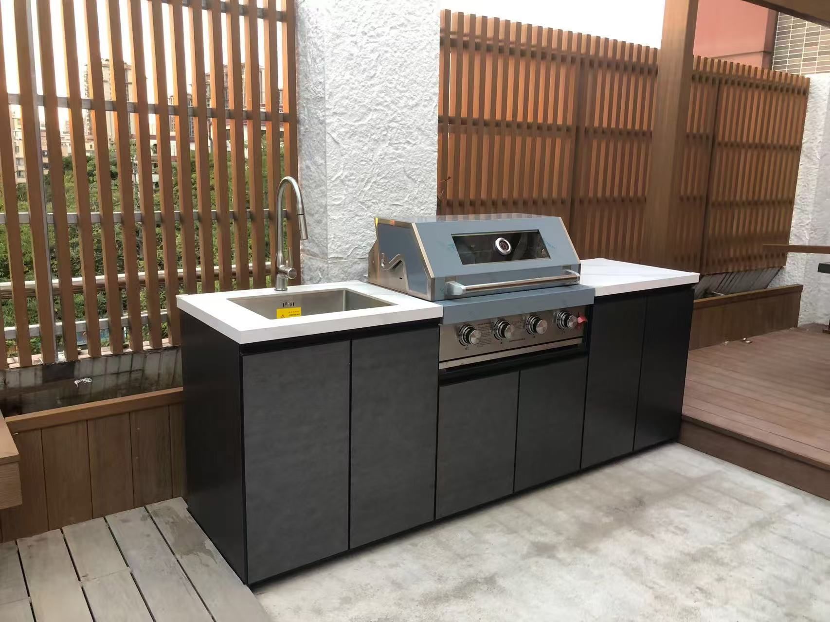 Outdoor kitchen with stainless grill, sink, and dark cabinets on a deck beside a wooden fence