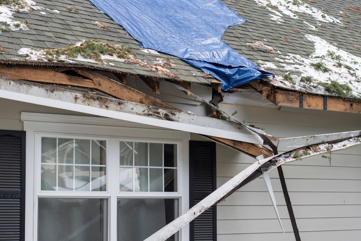House roof severely damaged; blue tarp covers some area; white siding, window visible.