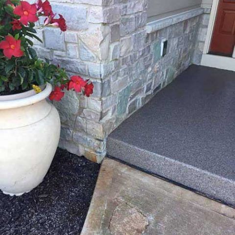Front porch with stone wall, gray carpeted step, red flowers in a white planter, and concrete walkway.