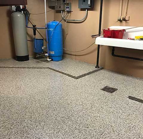 Utility room with blue water tanks, pipes, and a speckled floor drain beside a worktable.