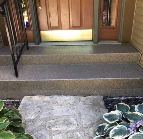 Front porch steps leading to a wooden front door, with a stone path and greenery in front.