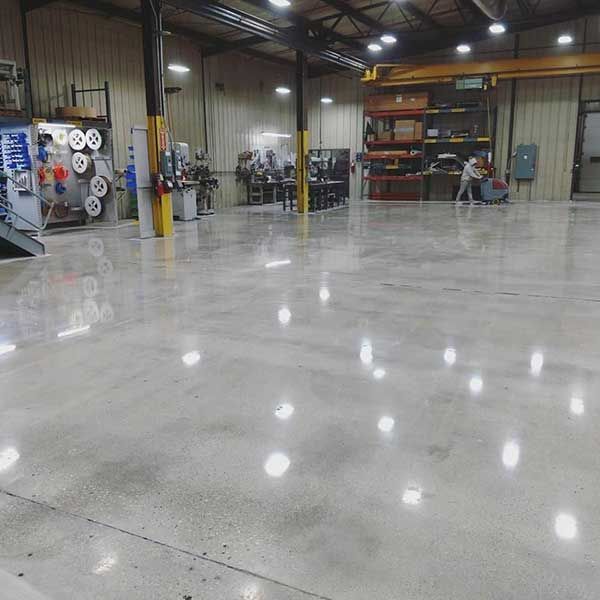 Spacious industrial workshop with polished concrete floor, metal shelving, equipment, and bright overhead lights.