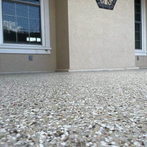 Low-angle view of a pebble-textured porch floor beneath a beige house wall and windows