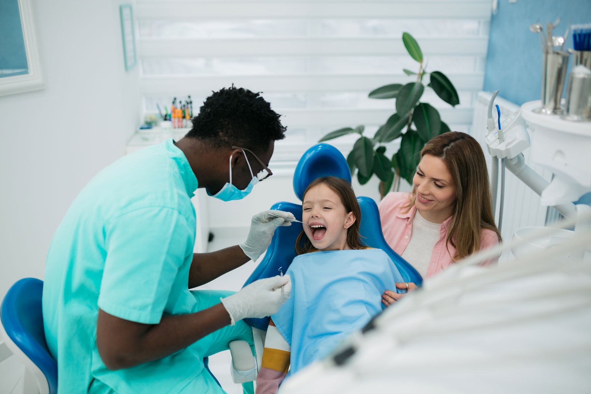 Family dentist examining a happy young girl’s teeth while her mother watches in a dental clinic.