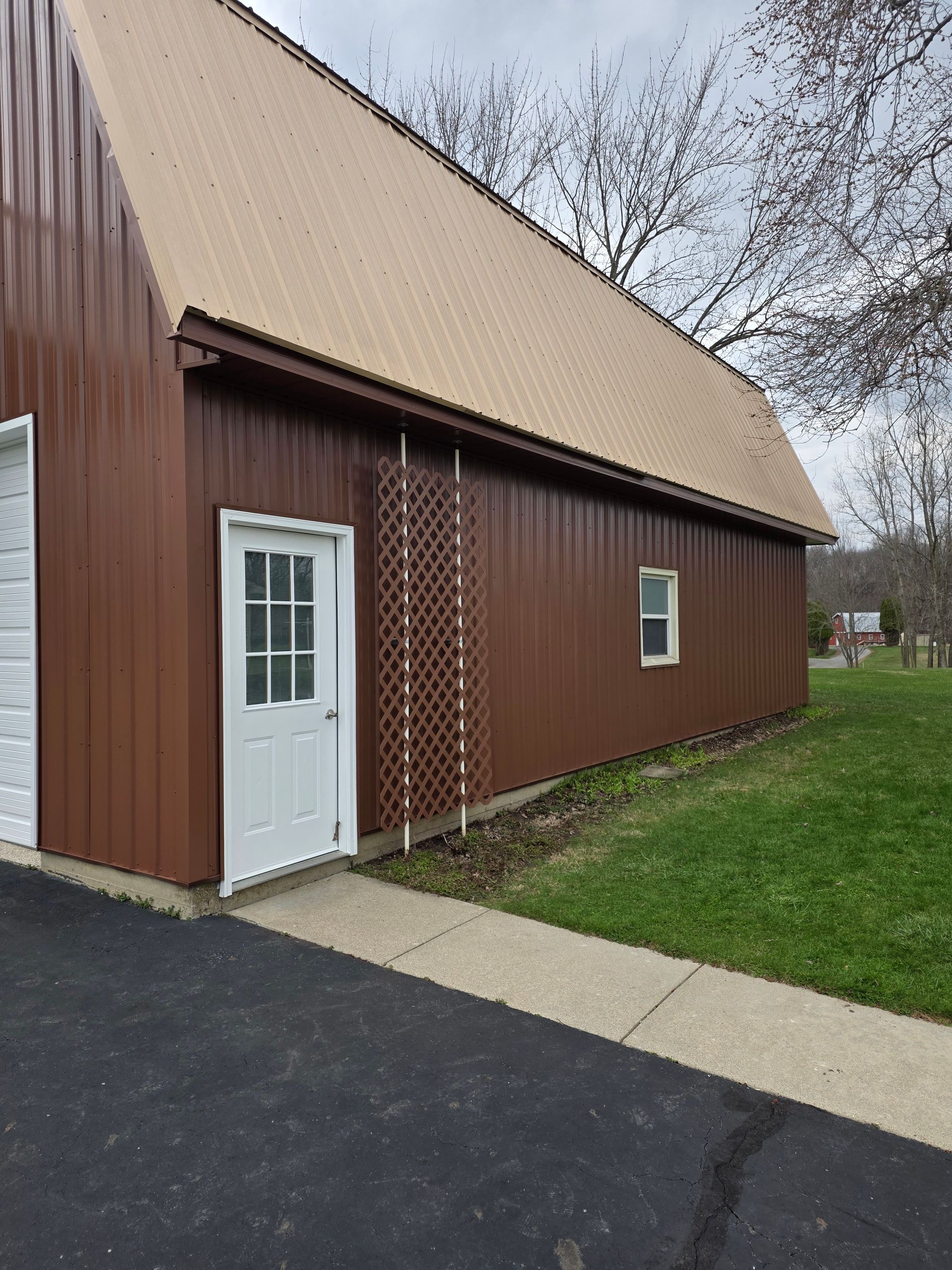 A brown metal-sided building with a beige roof, a white door, a trellis, and a single window next to an asphalt driveway.