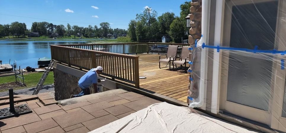 A person in a blue shirt works on a wooden deck by a lake, with a stone-paved patio and masked door in the foreground.