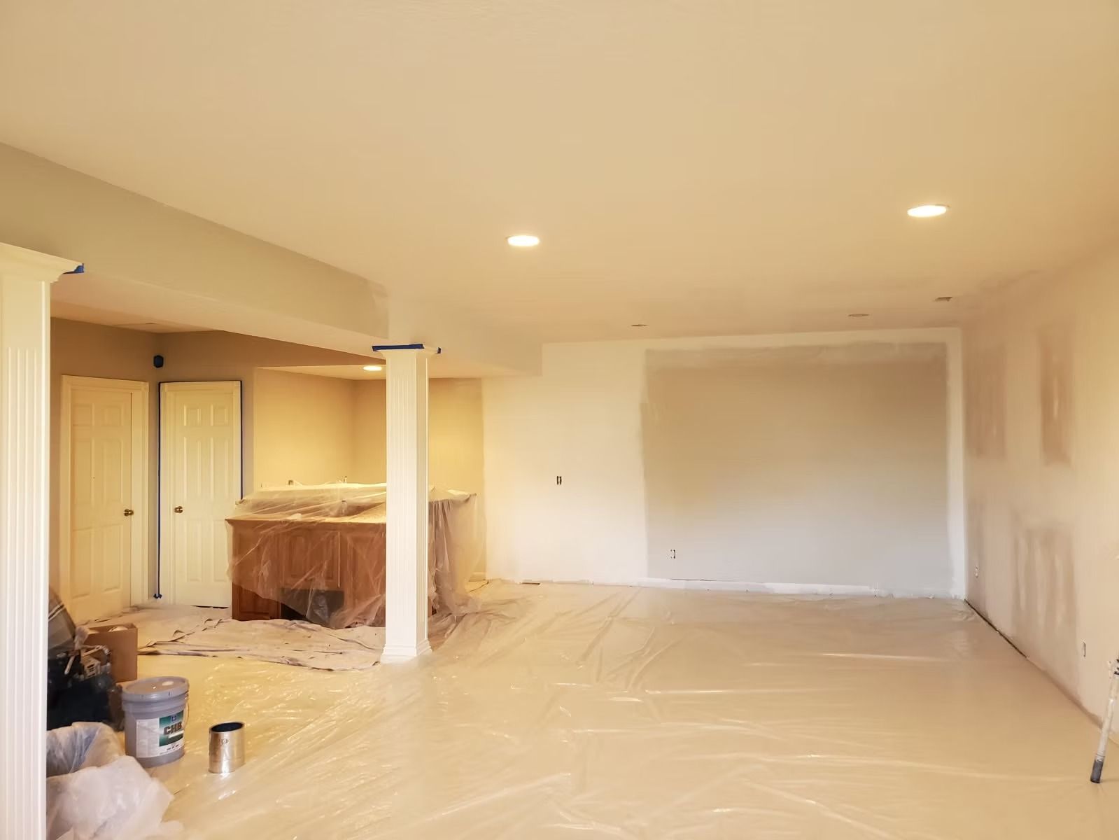An interior view of a room under renovation, featuring covered floors, white walls, support pillars, and recessed lighting.