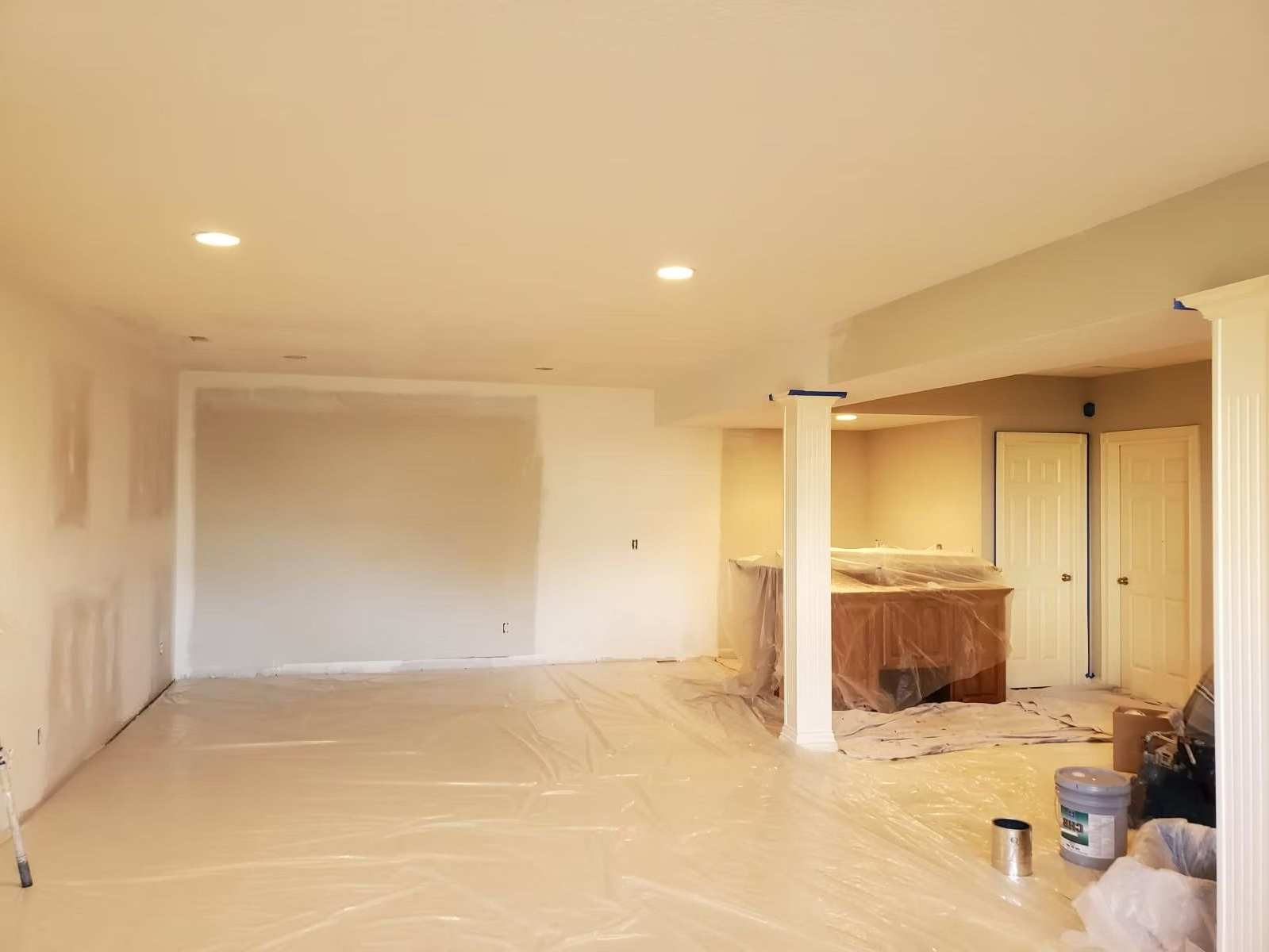 An unfinished room under renovation featuring white walls, a covered floor, two white columns, and recessed lighting.