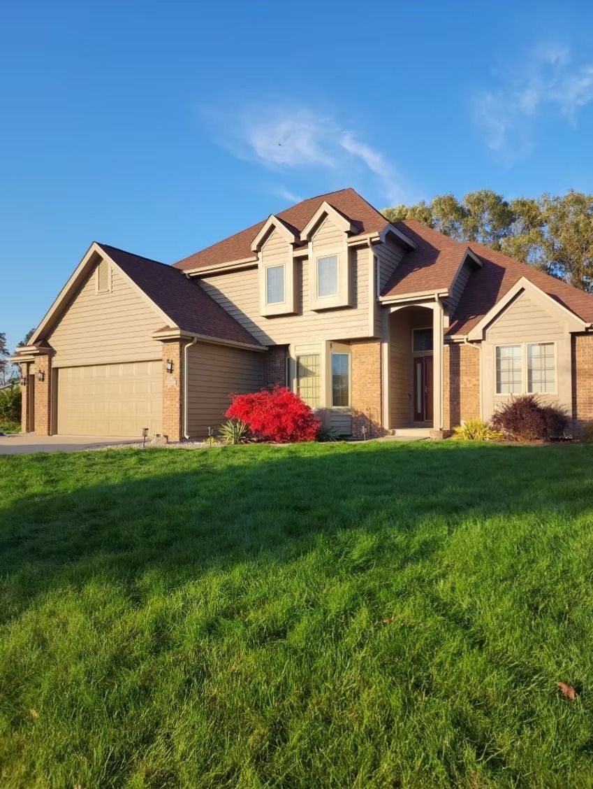 A two-story tan suburban house with a brown roof and attached garage, surrounded by a green lawn under a clear blue sky.