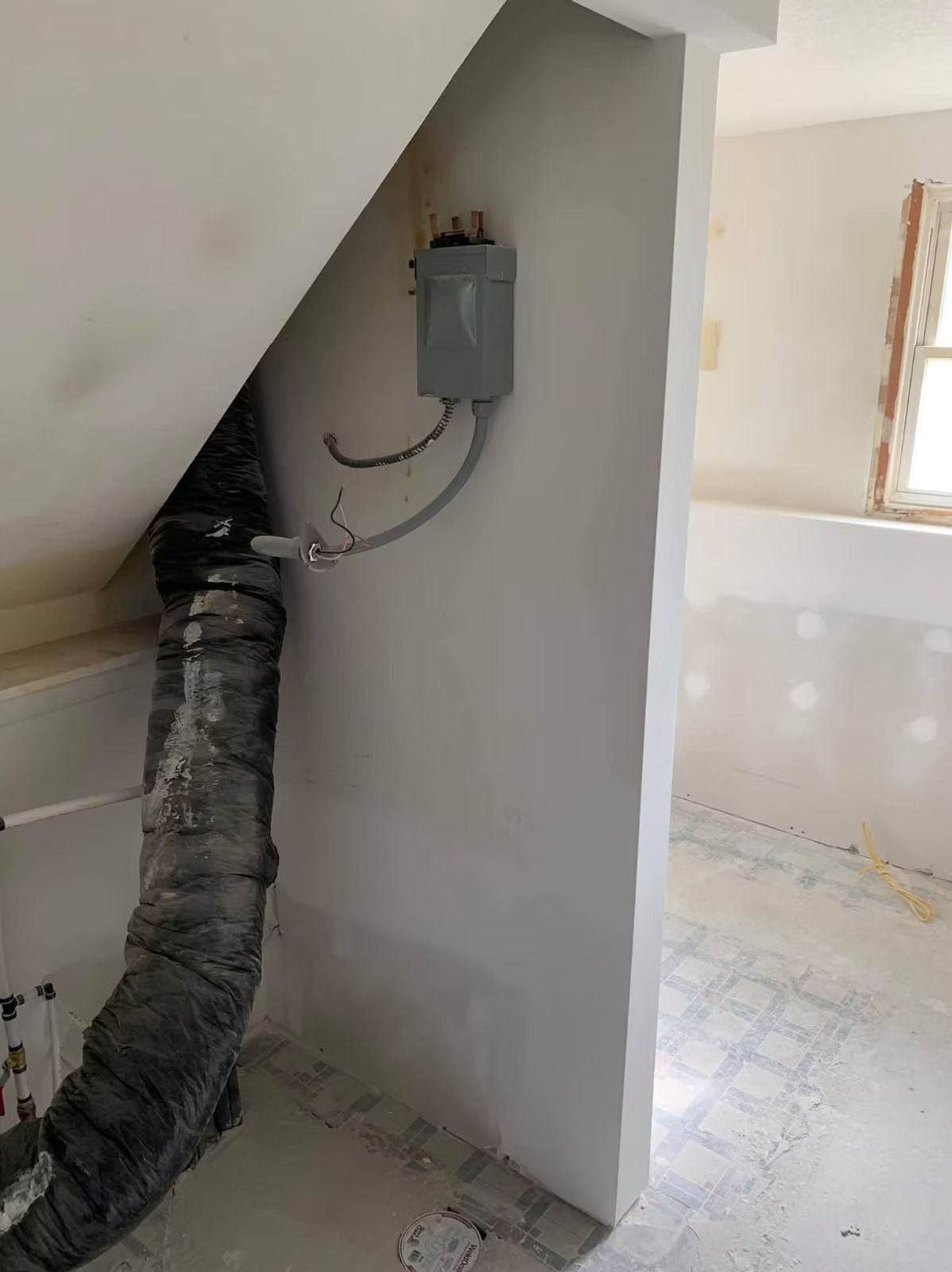 An unfinished room with a gray electrical disconnect box mounted on a white wall next to a large, insulated duct.