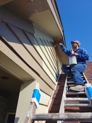 A person in a blue hoodie and jeans sits on a ladder, painting the exterior trim of a house under a clear blue sky.